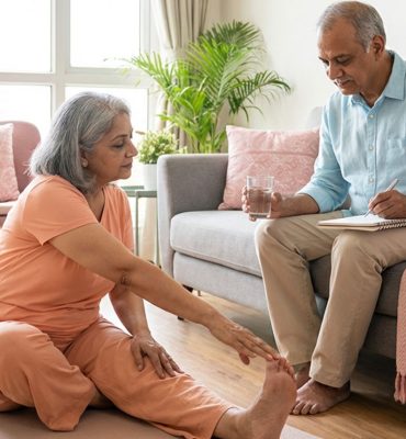 An elderly Indian man with white hair smiling warmly, sitting at a sunlit wooden table. He is surrounded by a laptop, a blood pressure monitor, a fresh green apple, and a glass of water, symbolizing a holistic and tech-savvy approach to senior wellness in 2026.