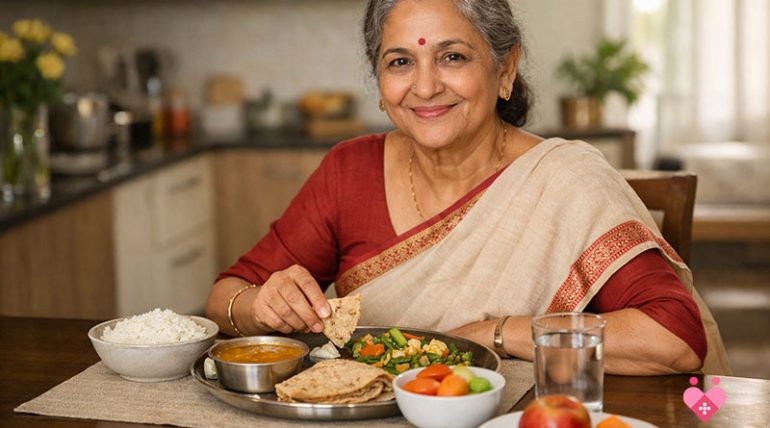 A smiling elderly Indian woman in a maroon and cream saree sitting at a dining table, enjoying a traditional meal. The plate includes roti, a bowl of dal, and sautéed vegetables, accompanied by a side of rice, fresh fruit, and a glass of water.
