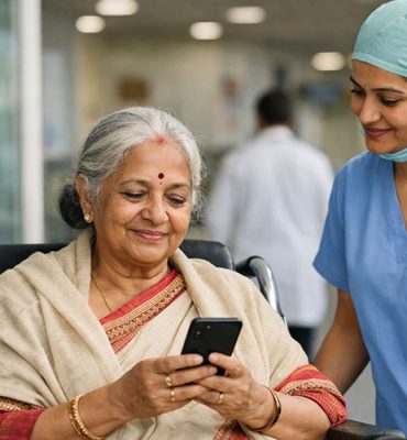 An elderly woman in a wheelchair smiling while looking at a smartphone, assisted by a healthcare professional in blue scrubs and a medical cap in a hospital setting.