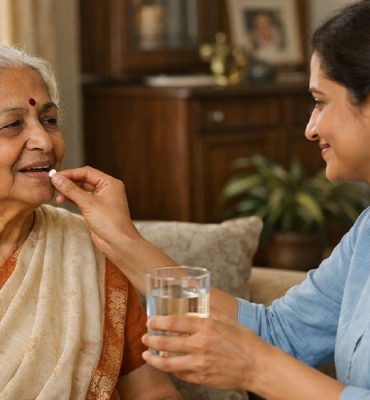 A young female caregiver assisting an elderly Indian woman with her medication by offering a pill and a glass of water in a comfortable home setting.